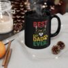 A black mug with the text 'BEST CAT DAD EVER' and a graphic of a cat wearing sunglasses and a paw print, set in a cozy, festive scene with a candle, pinecones, and fairy lights.