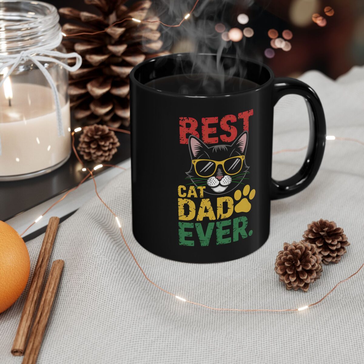 A black mug with the text 'BEST CAT DAD EVER' and a graphic of a cat wearing sunglasses and a paw print, set in a cozy, festive scene with a candle, pinecones, and fairy lights.