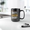 Black mug with text 'BEST DADDY EVER' in yellow and white, on a white desk with a notebook and pens.
