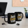 Two black mugs with the text 'BEST Daddy EVER' in white and yellow font, one in the foreground and another slightly behind it, placed on a wooden desk next to a laptop and a plant.