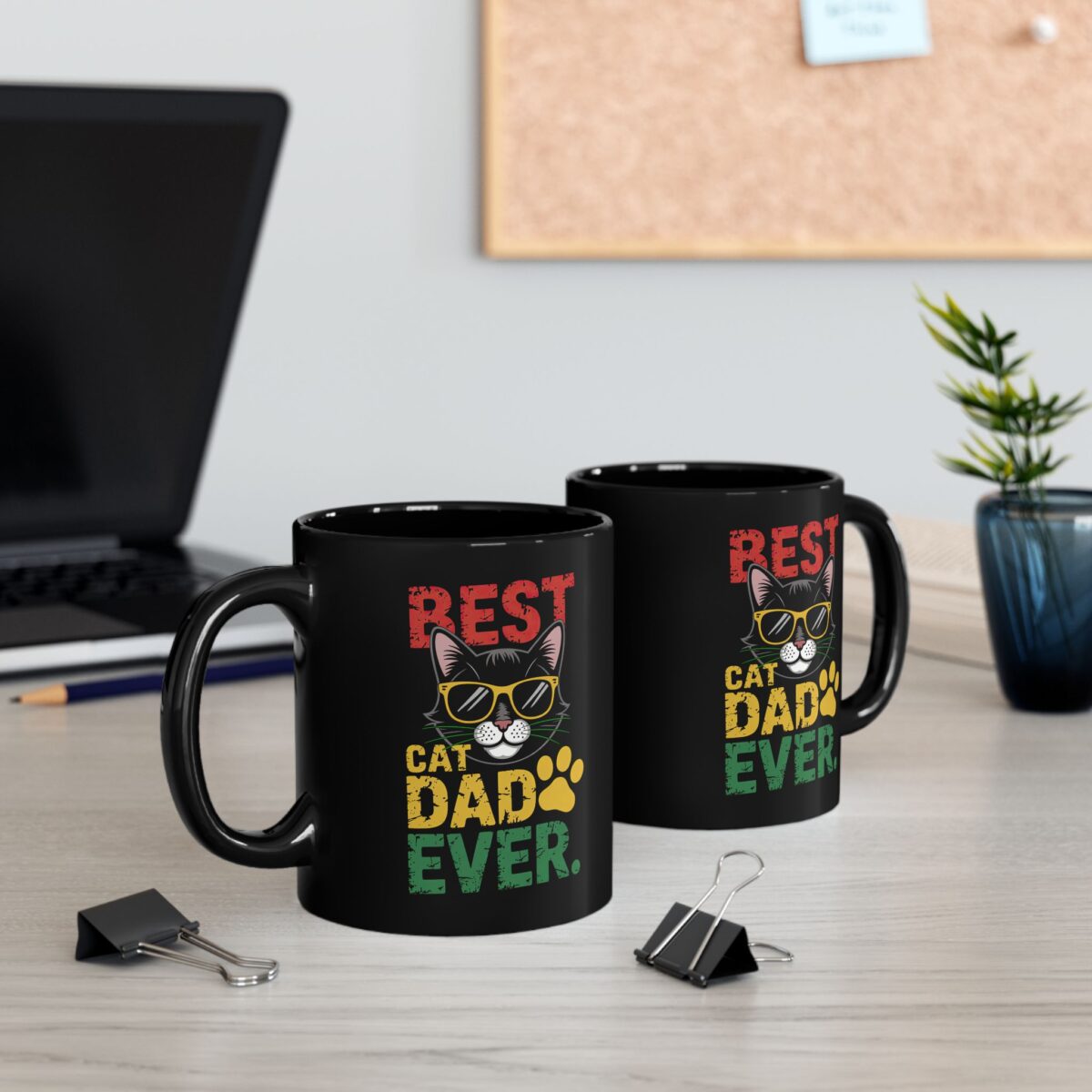 Two black mugs with a 'Best Cat Dad Ever' cat graphic on a desk.