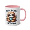 White ceramic mug with pink handle and rim, featuring a cute sloth illustration holding a polka dot mug. Text reads "BUSY DOING NOTHING".