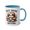 White ceramic mug with blue handle and interior, featuring a cartoon sloth holding a polka-dot cup and the phrase "BUSY DOING NOTHING".