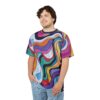 A young man with curly brown hair and a light beard is wearing a t-shirt with a vibrant, swirling, psychedelic pattern in colors including pink, orange, yellow, purple, blue, green, and black. He is also wearing light blue jeans and has his right hand in his pocket. He is looking off to the side with a slight smile.