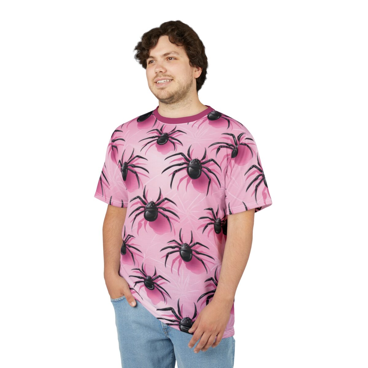A person with curly brown hair and a light beard smiles, wearing a pink t-shirt with black spider all-over print and light blue jeans.
