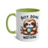 White ceramic coffee mug with a green handle and interior, featuring a cartoon sloth holding a blue polka dot mug.