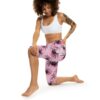A smiling woman with curly hair and tattoos kneels, wearing a white sports bra and pink capri leggings with black spider print.
