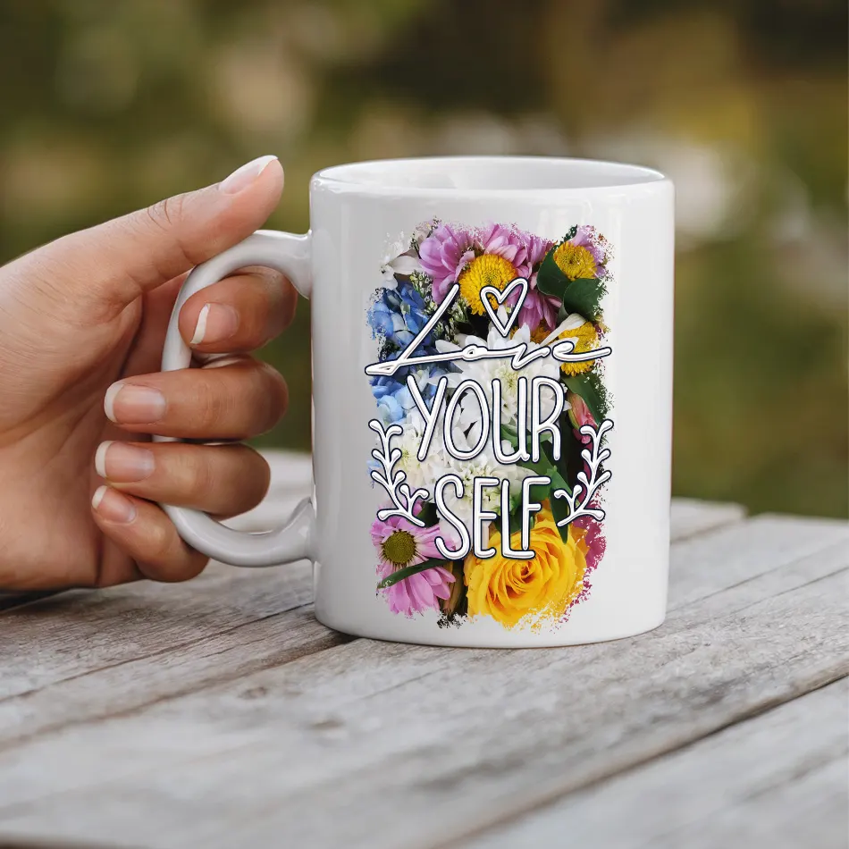 A white ceramic mug held by a hand, with colorful flowers and the text "Love Yourself"