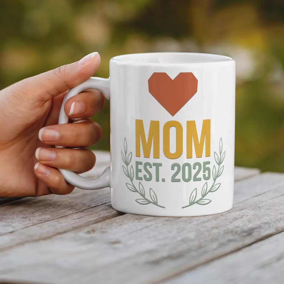 White ceramic mug with a geometric heart, "MOM EST. 2025" text, and leafy accents.