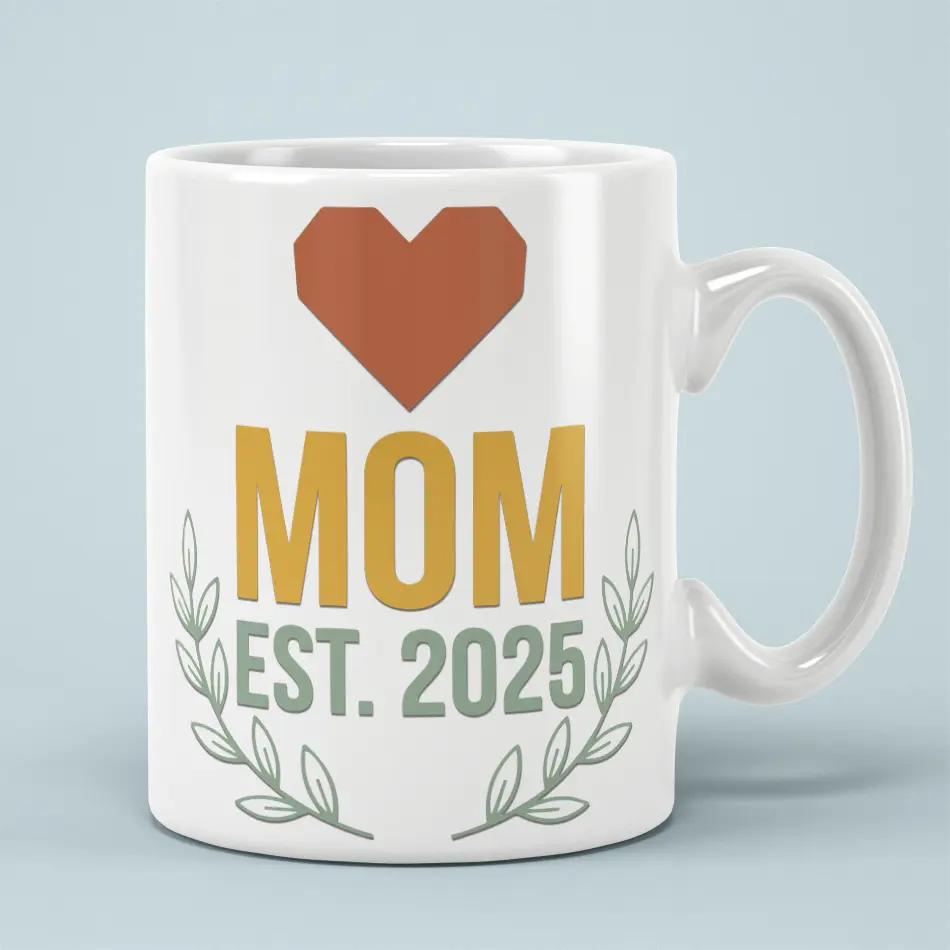White ceramic mug with a terracotta heart above "MOM EST. 2025" in mustard and green lettering, framed by leafy sprigs.