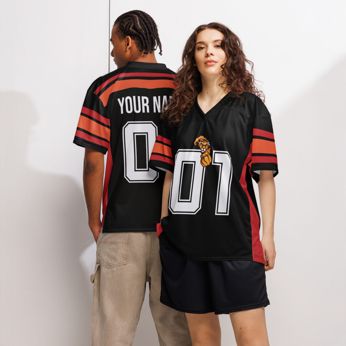 A man and a woman model wearing customizable black football jerseys with orange stripes on the sleeves and sides. The jerseys feature large white numbers and a small lion graphic. The back of the man's jersey shows 'YOUR NAME'.