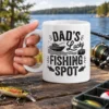 White ceramic coffee mug printed with black text reading Dad's Lucky Fishing Spot alongside boat and fish graphics.