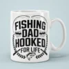 White ceramic coffee mug printed with bold black Fishing Dad Hooked For Life graphic artwork and bass fish icon.