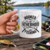 White ceramic mug printed with black text reading Hooked On Fatherhood And Fishing alongside fish graphics and fishing rods.