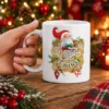 White ceramic mug featuring Santa Claus graphic with Santa Is Coming text and festive Christmas pine and berry accents.